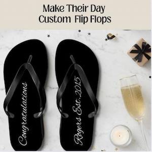 May include: Black flip-flops with white text that reads "Congratulations" and "Rogers Est. 2015". The image includes a champagne glass, a lit candle, and a wrapped gift. The text at the top says "Make Their Day Custom Flip Flops".