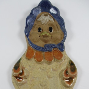 May include: A ceramic chicken-shaped dish with a blue bonnet, orange ruffle, and green and brown accents. The dish is glazed in a light brown color.