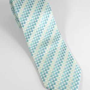 May include: A necktie featuring a repeating geometric pattern of small squares in shades of light blue, white, and pale green. The fabric has a textured appearance with a pointed tip and a folded top section, displaying a consistent grid design.