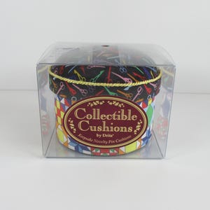 May include: A round pin cushion in a clear plastic box. The lid features a black fabric with a colorful scissor pattern and gold trim. The base displays a multi-colored geometric quilt pattern. The label reads Collectible Cushions by Dritz.