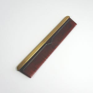 May include: A vintage comb with a dark red body and a gold-colored metal trim. The comb has fine teeth and a rectangular shape. The gold trim runs along the top edge of the comb, adding a touch of elegance to the design.