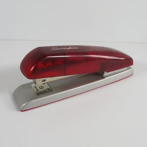 May include: A Swingline stapler featuring a translucent deep red plastic top cover and a solid silver-colored metal base. The brand name Swingline is printed in white script on the top of the red casing, which sits above a metallic stapling mechanism.