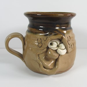 May include: A tan ceramic mug with a dark brown glazed rim and a sculpted face on the front. The face features two white protruding eyes and a wide, open mouth. The mug has a single curved handle on the left side and a smooth, glossy finish.