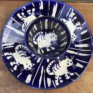 May include: A deep blue ceramic serving set consisting of a large round platter and a smaller matching bowl nested in the center. Both pieces feature a hand-painted white cat motif with striped patterns and vertical white brushstroke accents.