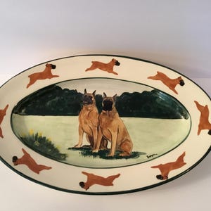 May include: Oval ceramic serving platter with a cream-colored background. The platter features a painted scene of two brown Boxer dogs sitting in a field, surrounded by running Boxer dog silhouettes and a green border.