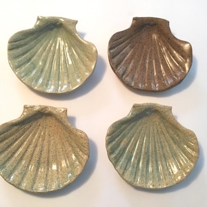 May include: Four ceramic dishes shaped like scallop shells. One dish is a solid light green, one is a solid brown, and two are a speckled light green and tan color. Each dish features a ribbed texture radiating from the hinge to the edge.
