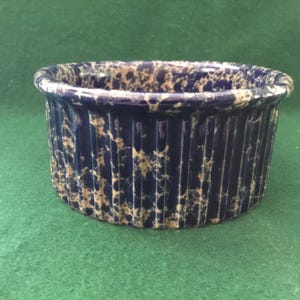 May include: A round ceramic ramekin featuring a deep blue base color with a speckled cream-colored pattern. The exterior of the dish has a vertical ribbed texture, and the rim is smooth and rounded, creating a consistent circular shape.