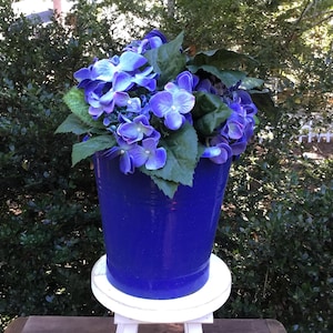 May include: A vibrant blue flower pot filled with blue and purple hydrangeas and green leaves. The pot sits on a small white stand, with a lush green background.