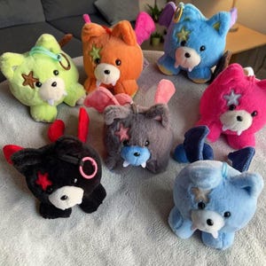 May include: A selection of colourful plush toys resembling bears, in shades of green, orange, blue, pink, grey, and black. Each has distinctive elements such as star patches, wings, and fangs. Some have keyrings attached.