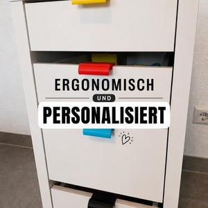 Könnte beinhalten: Ein weißer Schrank mit vier Schubladen, jede mit einem unterschiedlich farbigen Griff: gelb, rot, blau und schwarz. Die mittlere Schublade trägt den Text "ERGONOMISCH UND PERSONALISIERT" in einem schwarz-weißen Banner. Ein kleines Herz ist auf der unteren Schublade gezeichnet.