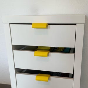Könnte beinhalten: Ein weißer Schrank mit vier Schubladen und leuchtend gelben Griffen. Der Schrank ist rechteckig und steht auf kleinen schwarzen Rollen. Die Schubladen sind geschlossen, vor einer weißen Wand.