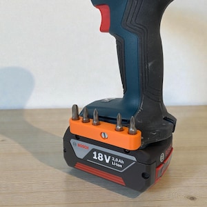 Könnte beinhalten: Ein kabelloser Bosch-Bohrer mit einem schwarzen und teal-farbenen Gehäuse, roten Akzenten und einem orangefarbenen Bit-Halter. Der Bohrer ist an einem schwarzen Akku mit dem Text "18V 3.0Ah Li-Ion" befestigt. Mehrere Bohrer sind in den orangefarbenen Halter eingesetzt.
