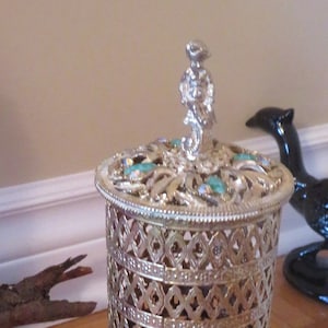 Pode incluir: Um recipiente decorativo de metal prateado com um design de filigrana e uma parte superior adornada com joias. O recipiente tem uma base redonda e um corpo cilíndrico. A parte superior do recipiente apresenta um remate decorativo.