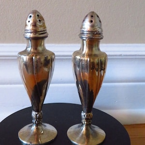 Puede incluir: Un par de saleros y pimenteros de plata con un diseño acanalado y una parte superior decorativa. Los saleros están sobre una superficie negra.