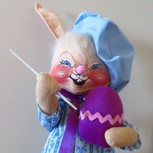 Può includere: Un coniglio di Pasqua di peluche che indossa un cappello blu e una camicia a righe blu e bianche. Il coniglio tiene un uovo di Pasqua viola e un pennello bianco.