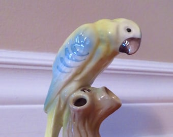 Puede incluir: Una figurita de cerámica de un loro amarillo y azul posado en un tocón de árbol. El loro tiene un ala azul claro y un pico marrón. El tocón del árbol es de color marrón claro y tiene un pequeño agujero en la parte superior.