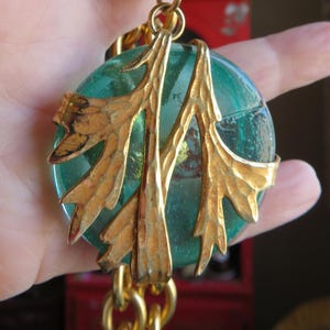 May include: A gold-toned necklace featuring a large, round, teal-colored pendant. The pendant is accented with gold-colored leaf-like embellishments. The necklace has a chunky chain design.