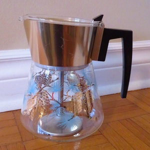 Puede incluir: Una cafetera de vidrio vintage con una tapa de metal dorada y un asa negra. La cafetera tiene un diseño de piñas y árboles de hoja perenne.