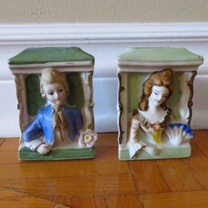 May include: Two vintage porcelain figurines, a man and a woman, each framed in a green and cream-colored structure. The man wears a blue jacket, and the woman has a dress with gold accents. The figurines are on a wooden surface.