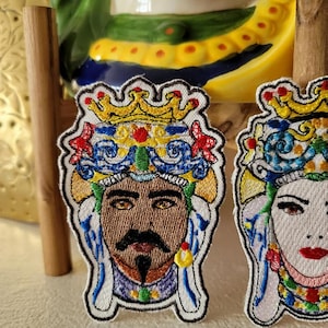 May include: Two embroidered patches featuring colorful, stylized portraits of people wearing crowns. The patches are white with blue, yellow, red, and green embroidery. The person on the left has brown skin and a black beard. The person on the right has white skin and red lips.