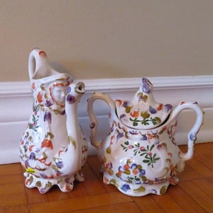 Puede incluir: Dos piezas decorativas de cerámica blanca. Una es una jarra con diseño de rostro y un pico curvo. La otra es una tetera con tapa y asas curvas. Ambas están adornadas con coloridos estampados florales en naranja, morado y verde.