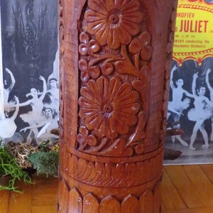 Puede incluir: Un jarrón de madera con intrincados tallados de flores y hojas. El jarrón es marrón y tiene una forma cilíndrica.