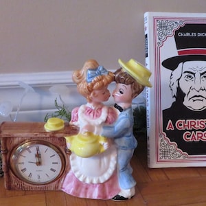 May include: A vintage ceramic clock figurine of a couple kissing. The clock is shaped like a wooden house with a yellow teapot and a white clock face. The couple are dressed in blue and pink with yellow hats. The clock is next to a book titled "A Christmas Carol" by Charles Dickens.