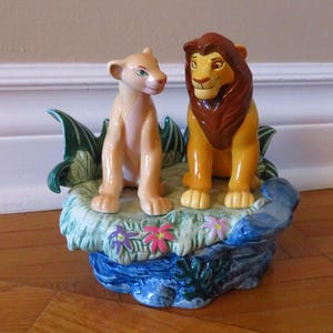 May include: A ceramic figurine of two lion characters, a male and a female, on a decorative base with green foliage, blue water, and colourful flowers. The lions are in shades of tan, brown, and yellow.