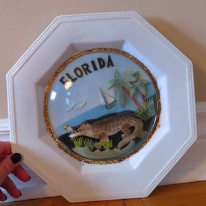 Puede incluir: Plato decorativo octogonal blanco con una imagen central de Florida. La imagen presenta un caimán detallado, palmeras, un velero y la palabra "FLORIDA" en letras negras. La imagen está rodeada por un borde dorado con cuentas.