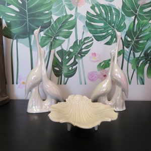 Puede incluir: Tres figuritas de pájaros de cerámica blanca con detalles iridiscentes, de pie sobre un plato de cerámica blanca con borde festoneado y cuatro patas. Los pájaros están dispuestos de forma simétrica, con un pájaro en el centro y dos pájaros a cada lado. El fondo es una pared blanca con un patrón floral verde y rosa.