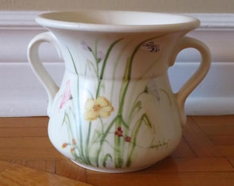 May include: Cream-colored ceramic vase with two handles and floral design. The vase features hand-painted yellow, pink, and purple flowers with green stems and leaves. The vase has a wide opening and sits on a wooden surface.