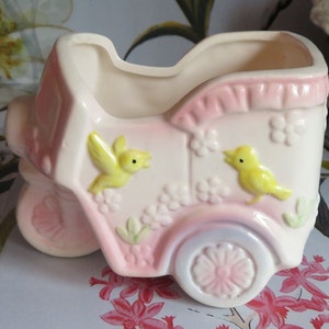 May include: A vintage ceramic baby carriage planter in soft pink and cream. The planter features embossed details, including yellow birds, flowers, and a decorative wheel. A charming piece for nurseries or vintage decor.