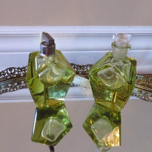 May include: Two vintage perfume bottles with a geometric design in a light green hue. One bottle has a silver-toned atomizer, the other a glass stopper. They sit on a mirrored tray with an ornate, silver-colored edge.