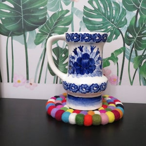 Puede incluir: Una jarra de cerámica azul y blanca con un diseño floral se encuentra sobre un posavasos de fieltro de colores. La jarra tiene un asa y una base ancha.