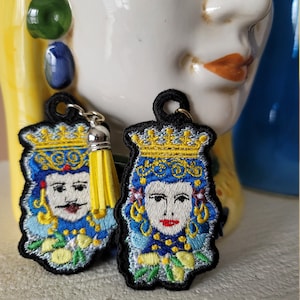 May include: A pair of embroidered earrings featuring a king and queen with crowns, yellow lemons, and blue and green details. The earrings have black backing and yellow tassels.