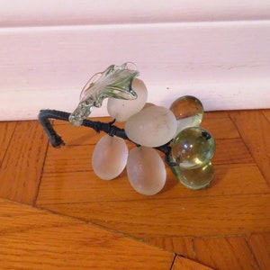May include: A glass grape cluster figurine with a green leaf on top. The grapes are frosted and clear, and the stem is black wire.