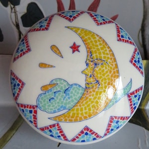 Könnte beinhalten: Eine weiße Keramik-Schmuckdose mit einem bunten Mosaikdesign. Der Deckel zeigt einen gelben Halbmond mit einem lächelnden Gesicht und einen kleinen roten Stern.