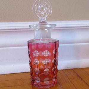 Peut inclure: Une bouteille en verre transparent avec un corps texturé rose et un bouchon en verre transparent. La bouteille a un design unique et orné avec un motif circulaire.
