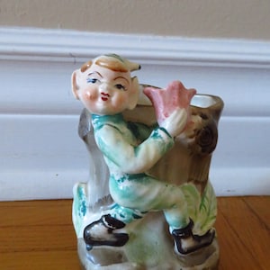 May include: A ceramic figurine of a whimsical elf holding a pink flower. The elf is wearing a green and blue outfit and is standing on a tree stump. The figurine is a vintage planter.
