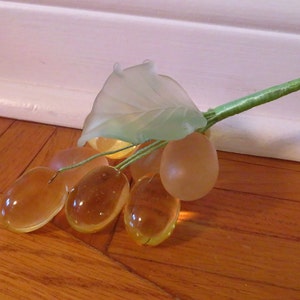 May include: A cluster of glass grapes with a green leaf. The grapes are a light amber color and the leaf is a light green.