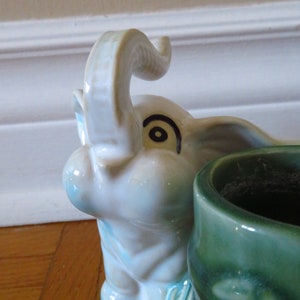 Op de afbeelding: Een keramische plantenpot in de vorm van een olifant met een groene plantenpot op zijn rug. De olifant is wit met blauwe en groene accenten en heeft een zwart oog met een swirl-ontwerp.