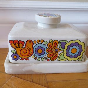 May include: White ceramic butter dish with a lid and a decorative floral and bird design in orange, yellow, blue, and purple. The lid has a small, round handle with a floral pattern.