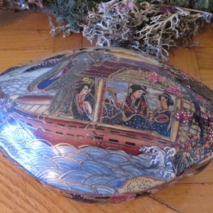 Puede incluir: Una caja de cerámica para baratijas con forma de concha con una escena pintada de mujeres japonesas en un barco sobre un fondo de olas azules y blancas.