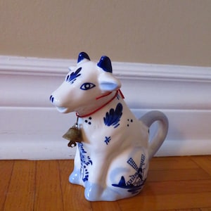 May include: A white and blue ceramic cow creamer with a bell around its neck. The cow creamer has blue accents and a windmill design. The creamer is sitting on a wooden surface.