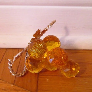 May include: A cluster of amber glass grapes with a gold wire stem. The grapes are faceted and have a shiny finish.
