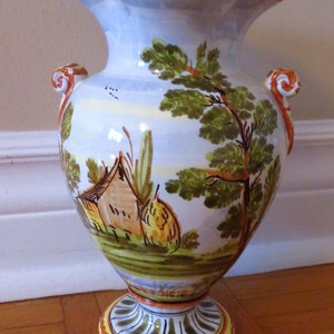 May include: A white ceramic vase with a scalloped rim and two scroll handles. The vase is decorated with a hand-painted scene of a house, trees, and a field. The vase has a yellow, orange, and green base.