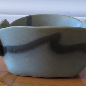 Puede incluir: Un cuenco de cerámica verde con un diseño abstracto ondulado en esmalte negro. El cuenco tiene una forma única y orgánica con una abertura ancha y redondeada y una base ligeramente curvada.