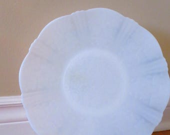 Könnte beinhalten: Ein weißer Milchglasteller mit einem gewellten Rand. Der Teller steht auf einer Holzoberfläche und wird von zwei durchsichtigen Plastikständern gehalten.