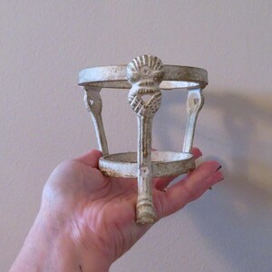 May include: A distressed white metal plant stand with a decorative seashell design. The stand has a circular top and base, connected by three curved legs and a central support. The stand is approximately 15 cm tall.