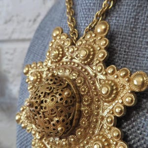 Op de afbeelding: Een goudkleurige statement ketting met een grote, sierlijke hanger. De hanger heeft een gedetailleerd, opengewerkt ontwerp met een centrale bol en stralende bloemachtige elementen. De ketting is een eenvoudige schakelstijl.